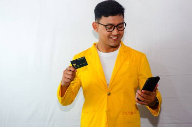 Sarı ceketli ve gözlüklü genç Endonezyalı adam, kredi kartı gösteriyor ve gülümserken akıllı telefon tutuyor.