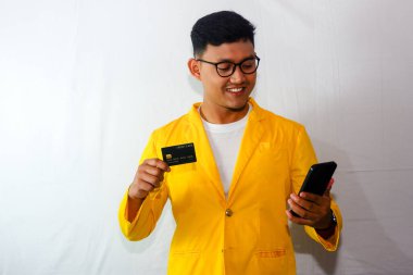 Sarı ceketli ve gözlüklü genç Endonezyalı adam, kredi kartı gösteriyor ve gülümserken akıllı telefon tutuyor.