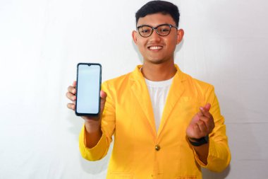 Endonezyalı genç adam sarı takım elbiseli ve gözlüklü, akıllı telefon boş ekranı gösteriyor ve sarangheo hareketi yapıyor.