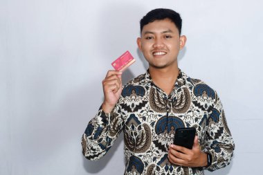 Genç Endonezyalı batik giyen sakin yüzlü kredi kartı ve cep telefonu tutan adam.