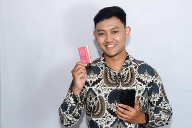 Genç Endonezyalı gülen yüzlü adam kredi kartı gösteriyor ve elinde cep telefonu tutuyor.
