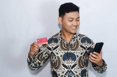 Genç Endonezyalı gülen yüzlü adam kredi kartını gösteriyor ve cep telefonuna bakıyor.