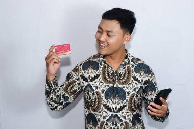 Genç Endonezyalı gülen yüzlü adam kredi kartına bakıyor ve elinde cep telefonu tutuyor.