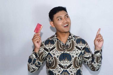 Genç Endonezyalı, gülen yüzlü, batik giyip kredi kartı gösteren ve...