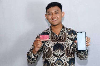 Genç Endonezyalı gülen yüzlü adam kredi kartını gösteriyor ve elinde boş ekranlı cep telefonu tutuyor.