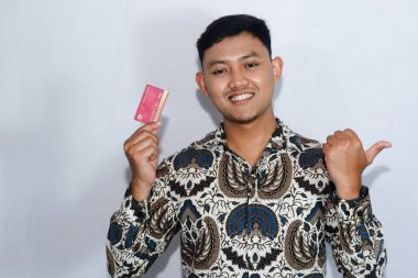 Genç Endonezyalı gülen yüzlü adam kredi kartını gösteriyor ve başparmağıyla yüzünü işaret ediyor.