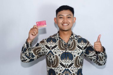 Genç Endonezyalı gülen yüzlü, batik giyen kredi kartı gösteren ve başparmağını kaldıran bir adam.