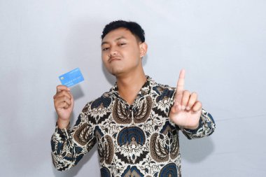 Sakin yüzlü batik giyen genç Endonezyalı adam kredi kartı gösteriyor ve dur işareti yapıyor
