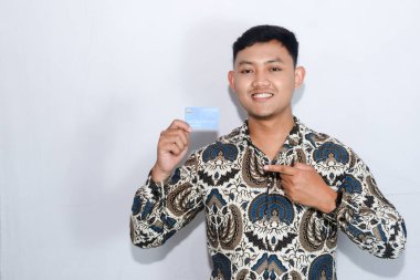 Genç Endonezyalı batik giyen, şaşırmış yüzlü kredi kartı gösteren ve kredi kartını işaret eden bir adam.