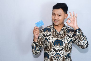 Genç Endonezyalı batik giyen, dost yüzlü kredi kartı gösteren ve iyi bir jest yapan bir adam.