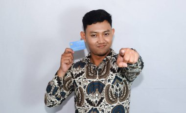 Dost yüzlü batik giyen genç Endonezyalı adam kredi kartı gösteriyor ve ileri işaret ediyor.