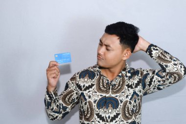 Genç Endonezyalı Batik giyen, kafası karışmış, kredi kartına bakan bir adam.