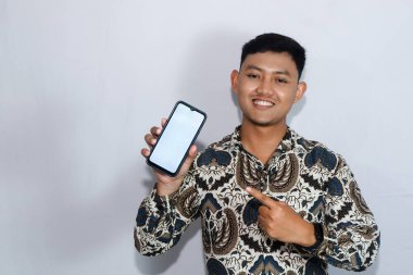 Batik giyen, sakin yüzlü genç Endonezyalı adam boş cep telefonu ekranını işaret ediyor.