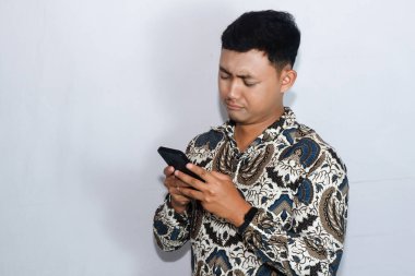 Genç Endonezyalı adam cep telefonuna bakarken üzgün yüzlü batik giyiyor.