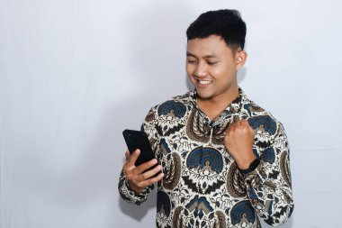 Batik giyen Endonezyalı genç adam cep telefonuna gülen yüzüyle bakıyor.