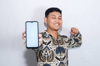 Batik giyen genç Endonezyalı adam boş cep telefonu ekranı gösteriyor ve mutlu bir yüz takınıyor.