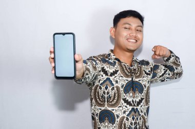 Batik giyen genç Endonezyalı adam boş cep telefonu ekranı gösteriyor ve mutlu bir yüz takınıyor.
