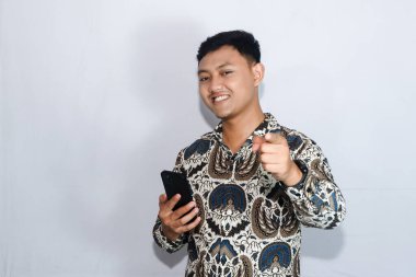 Genç Endonezyalı adam elinde cep telefonuyla gülümseyen ve mutlu yüzüyle ileri doğru işaret ediyor.