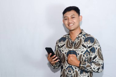 Dost canlısı Endonezyalı genç adam elinde cep telefonuyla batik takıyor ve başarıyı kutluyor.