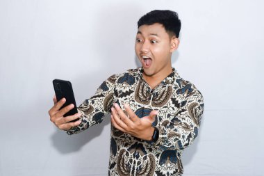 Elinde cep telefonu tutan, Batik giyen Endonezyalı genç bir adam şok oldu.