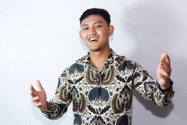 Batik giyen genç Endonezyalı adam gülümserken hoş geldin jesti yapıyor.