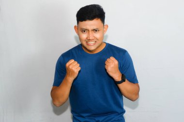 Asyalı mavi tişörtlü genç adam sinir bozucu hareketler yapıyor ve yumruğunu sıkıyor.