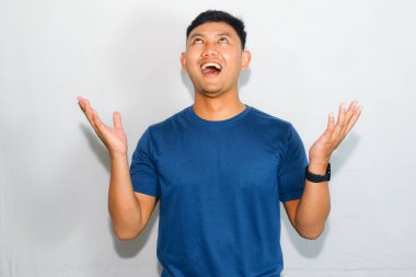 Mavi gömlekli genç Asyalı adam orta boy jest yapıyor ve ellerini kaldırıyor.