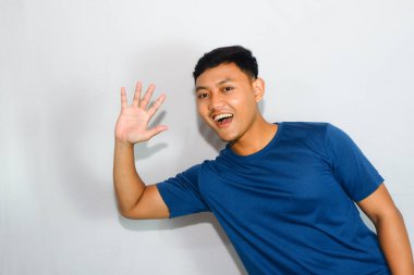 Mavi tişörtlü, mutlu yüzlü, selamlama hareketi yapan genç Asyalı bir adam.