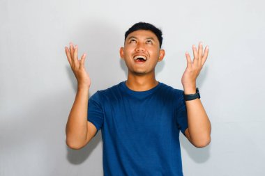 Mavi tişörtlü genç Asyalı adam, mutlu bir ifadeyle, el kaldırma hareketi yapıyor.