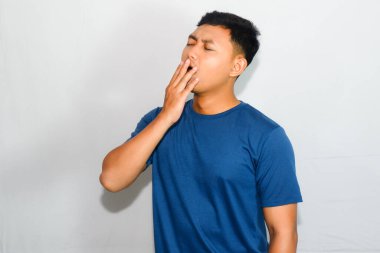 Mavi tişörtlü genç Asyalı adam uykulu olduğu için esniyor.