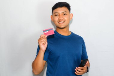 Cep telefonu tutarken kredi kartı gösteren mavi tişörtlü genç Asyalı adam.