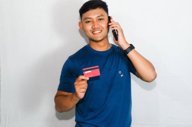 Mavi tişörtlü genç Asyalı adam cep telefonuyla konuşurken kredi kartı gösteriyor.