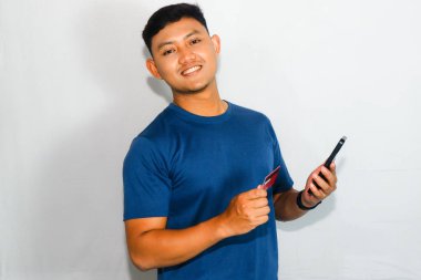 Cep telefonu tutarken kredi kartı gösteren mavi tişörtlü genç Asyalı adam.