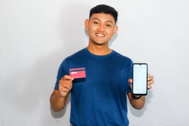 Mavi tişörtlü genç Asyalı adam kredi kartı ve boş cep telefonu ekranı gösteriyor.