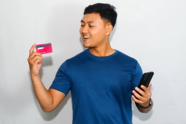 Kredi kartına bakarken mavi tişörtlü genç Asyalı adam kredi kartını gösteriyor.