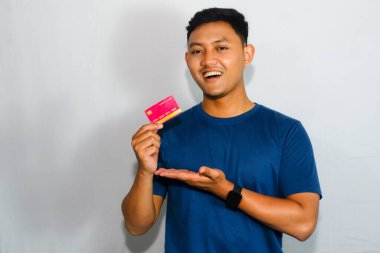 Mavi tişörtlü genç Asyalı adam gülümseyen yüzüyle kredi kartı gösteriyor.