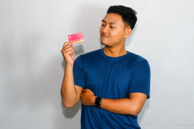 Mavi gömlekli genç Asyalı adam kredi kartını gösteriyor ve kollarını kavuşturuyor.