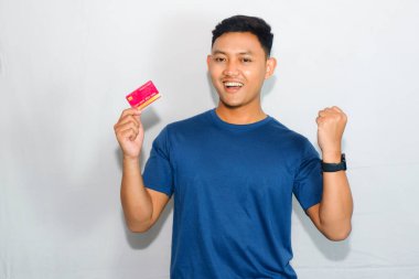 Mavi gömlekli genç Asyalı adam kredi kartı gösteriyor ve zaferi kutlamak için jest yapıyor.
