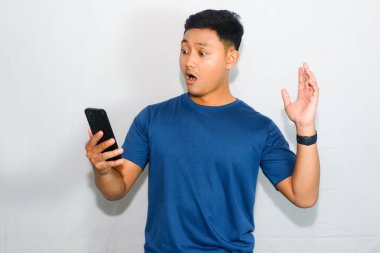 Dost canlısı mavi tişörtlü genç Endonezyalı adam elinde akıllı telefon tutuyor ve sürpriz jest yapıyor.