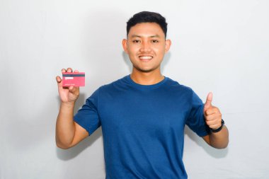 Mavi tişörtlü genç Asyalı adam kredi kartı gösteriyor ve düşünüyor ya da kafası karışık hareket ediyor.