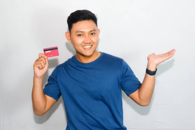 Mavi tişörtlü genç Asyalı adam kredi kartı gösteriyor ve kenarda işaret işareti yapıyor.