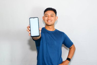 Dost canlısı genç Endonezyalı bir adam mavi tişört giyiyor, akıllı telefonunu boş bir ekranla ve tarafsız veya davetkar bir ifadeyle gösteriyor