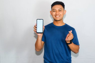 Dost canlısı genç Endonezyalı adam mavi tişört giyiyor, akıllı telefonunu boş bir ekranla gösteriyor ve 