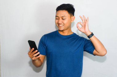 Dost canlısı genç Endonezyalı adam mavi tişörtlü, akıllı telefonuna bakarken olumlu bir ifadeyle 