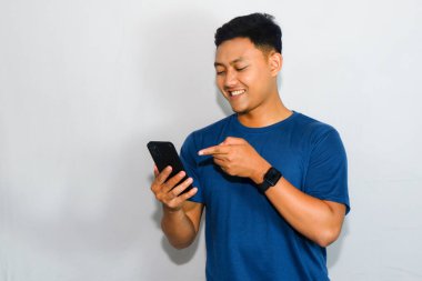 Dost canlısı genç Endonezyalı bir adam mavi bir tişört giyiyor, akıllı telefonunu tutuyor ve çekici bir ifadeyle işaret ediyor..