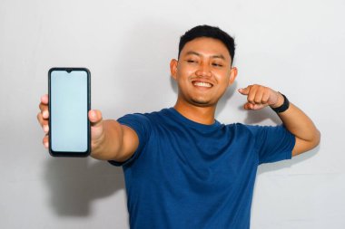 Dost canlısı genç Endonezyalı bir adam mavi tişört giyiyor, akıllı telefonunu boş bir ekranla gösteriyor ve tek eliyle ileri doğru işaret ediyor.