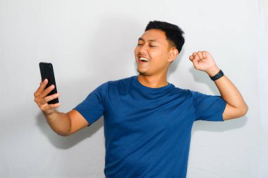 Dost canlısı genç Endonezyalı adam mavi tişört giyiyor, elinde akıllı telefonu var ve neşeli bir ifadeyle zafer kutlaması jesti yapıyor.