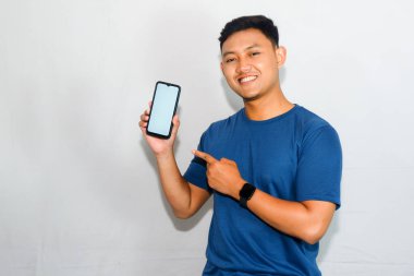 Dost canlısı genç Endonezyalı adam mavi tişört giyiyor, akıllı telefonunu gösteriyor ve tek eliyle işaret ediyor.