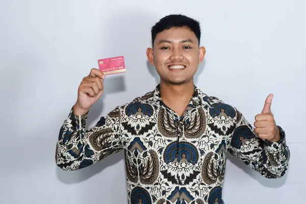 Genç Endonezyalı gülen yüzlü, batik giyen kredi kartı gösteren ve başparmağını kaldıran bir adam.