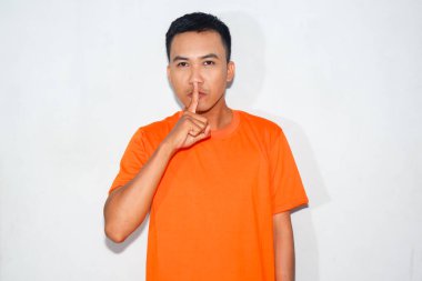 Turuncu tişört giyen heyecanlı bir Asyalı ciddi bir ifadeyle parmağını kullanarak ağzını kapatıyor.